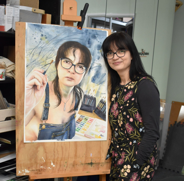 Woman standing next to a painting of herself on an easel in an art studio.